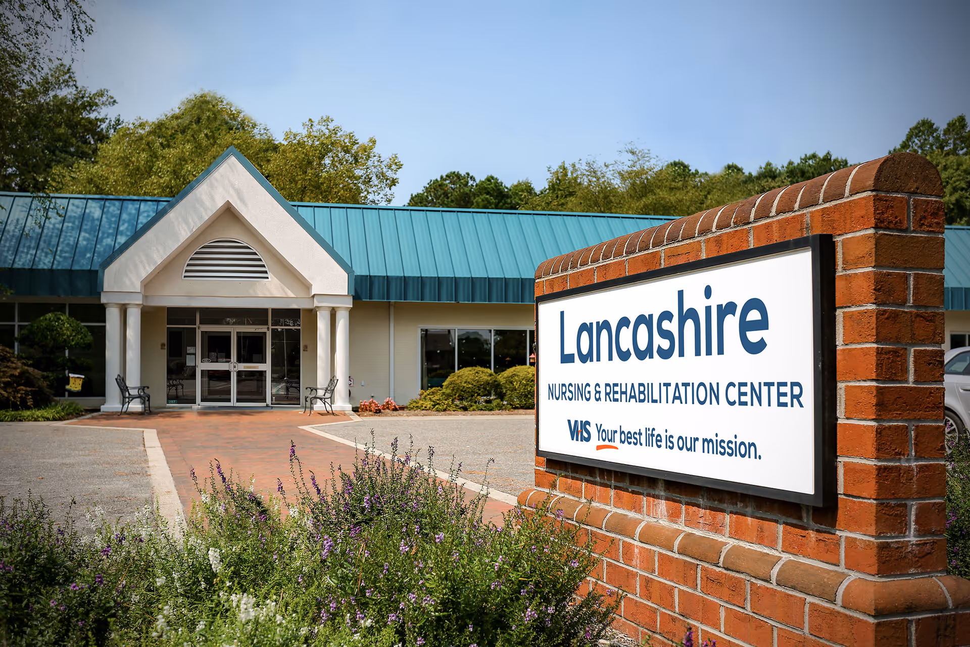 Exterior view of Lancashire Nursing & Rehabilitation Center showing the entrance with a peaked roof and columns, a brick sign in the foreground with the facility name and slogan, surrounded by greenery and flowers under a clear blue sky.