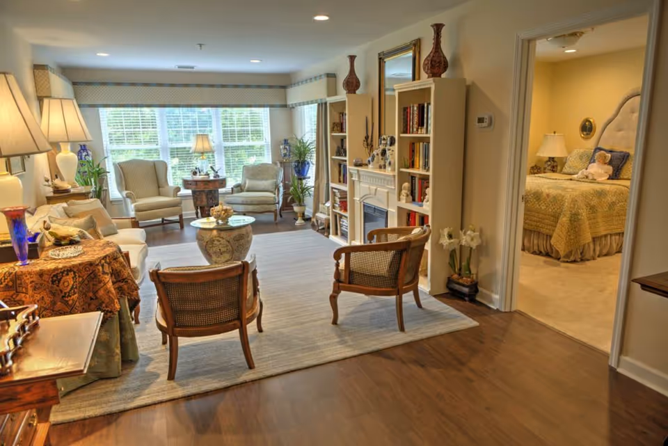 A cozy living room in a retirement community with wooden flooring, a large area rug, two wooden chairs, a sofa, and two armchairs near a large window. The room features a fireplace with bookshelves on either side, decorative vases, and lamps. An adjacent bedroom with a bed, nightstand, and lamp is visible through an open doorway.