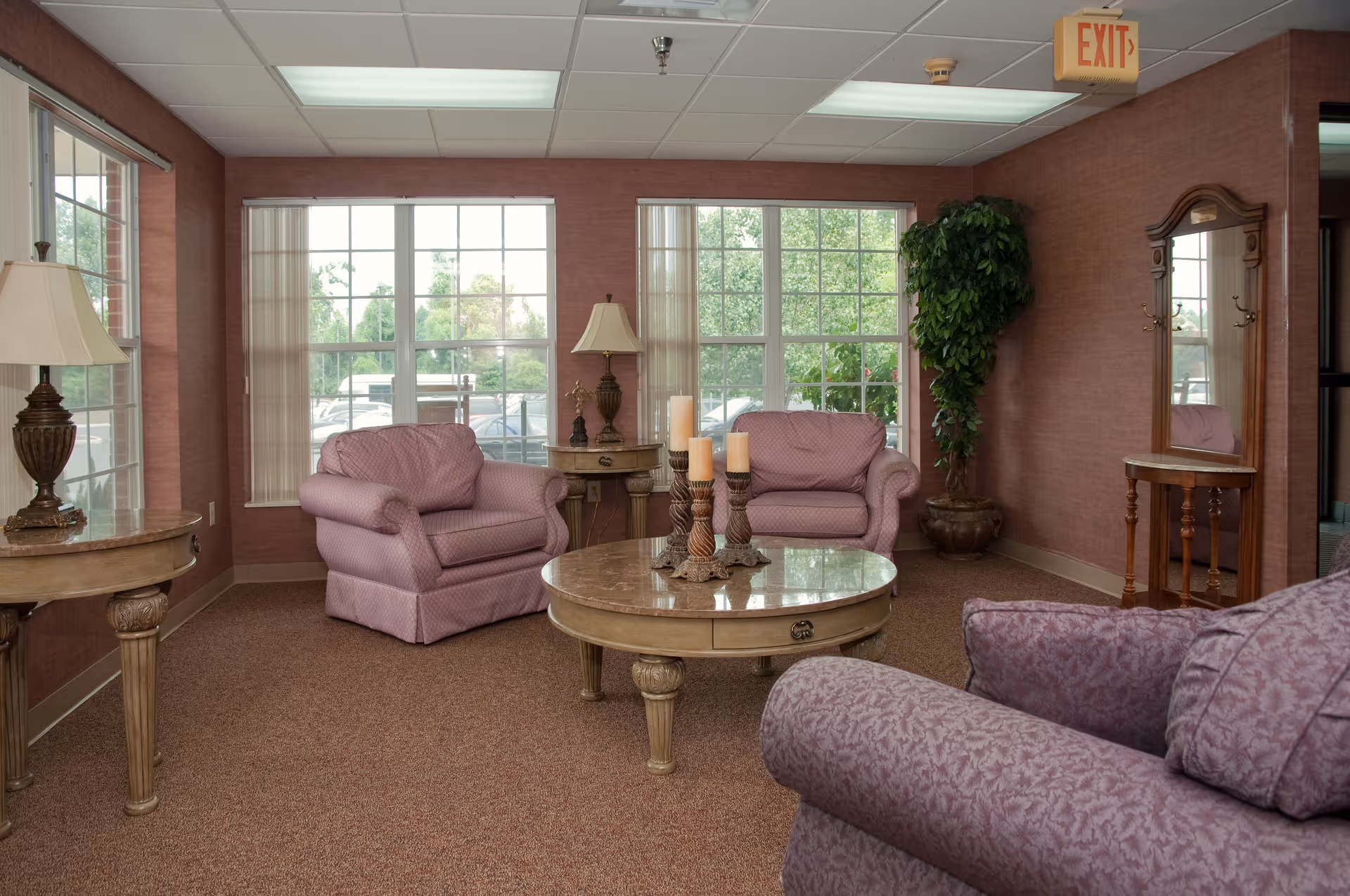 A cozy sitting area in a nursing and rehabilitation center with three upholstered armchairs in a soft purple pattern arranged around a round wooden coffee table with decorative candles. The room has large windows with vertical blinds, two table lamps on side tables, a tall potted plant in the corner, and a wooden hall tree with a mirror and hooks. The walls are covered in a textured reddish wallpaper and the floor is carpeted.