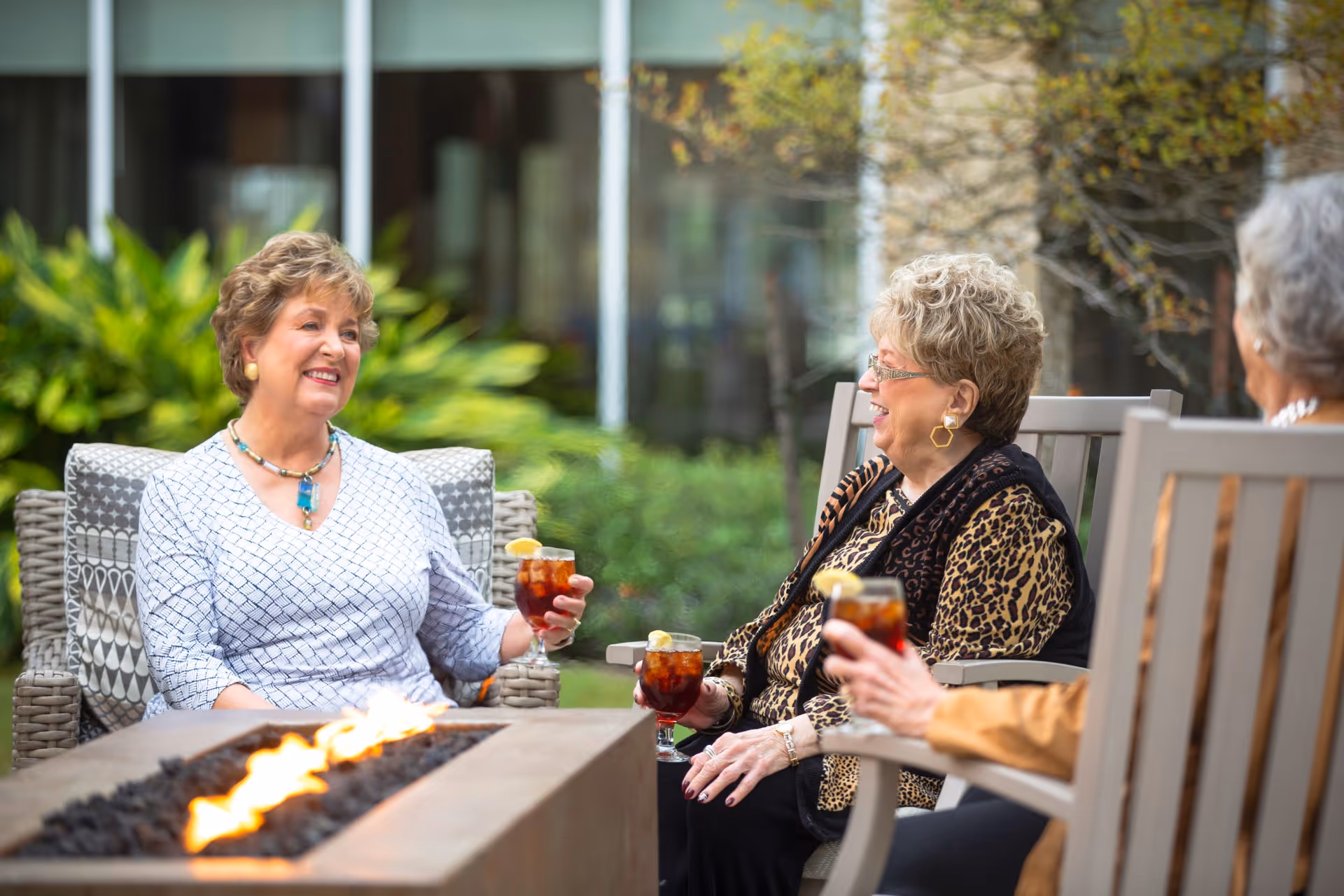 Three elderly women sitting outdoors around a rectangular fire pit, enjoying drinks with lemon garnishes and engaging in conversation. They are seated on cushioned chairs with greenery and a building in the background.