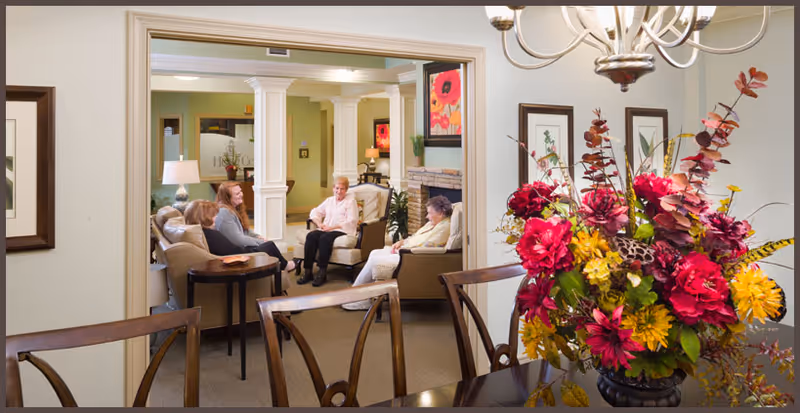 A cozy living room area in a senior living facility with four elderly women sitting and chatting on comfortable chairs and sofas. The room features warm lighting, framed artwork on the walls, a fireplace, and a large floral arrangement on a dining table in the foreground.
