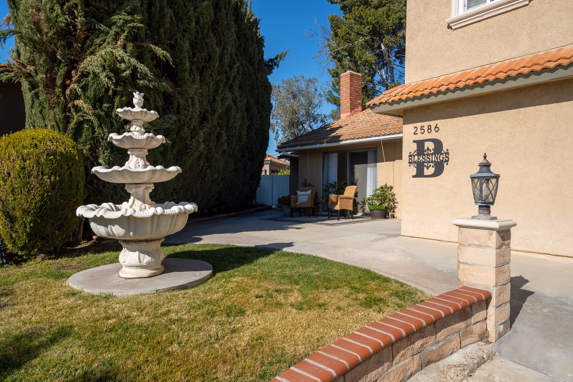 Outdoor view of a senior care facility with a multi-tiered white fountain on a grassy area, a beige building with a tiled roof, two chairs with cushions on a small patio, and the sign '2586 B Blessings' on the building wall.