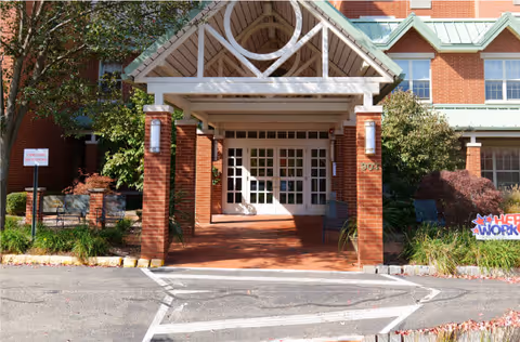 Entrance to a senior living facility with a covered porch supported by brick pillars and white wooden beams. The building is made of red brick with large windows and green metal roofing. There are plants and bushes on either side of the entrance and a sign on the right side that reads 'THE WORK'.