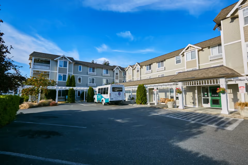Exterior view of La Conner Retirement Inn, a three-story building with beige siding and white trim. There is a parking area in front with a shuttle bus parked near the entrance. The entrance has a covered walkway with white pillars and hanging flower baskets. The sky is clear and blue.