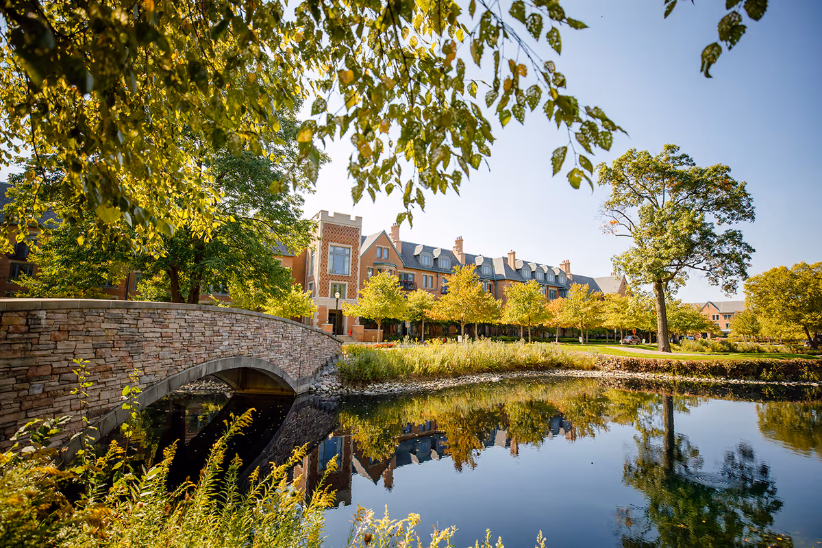 A stone footbridge and pond in front of a tree-lined senior living building reflected in the water.
