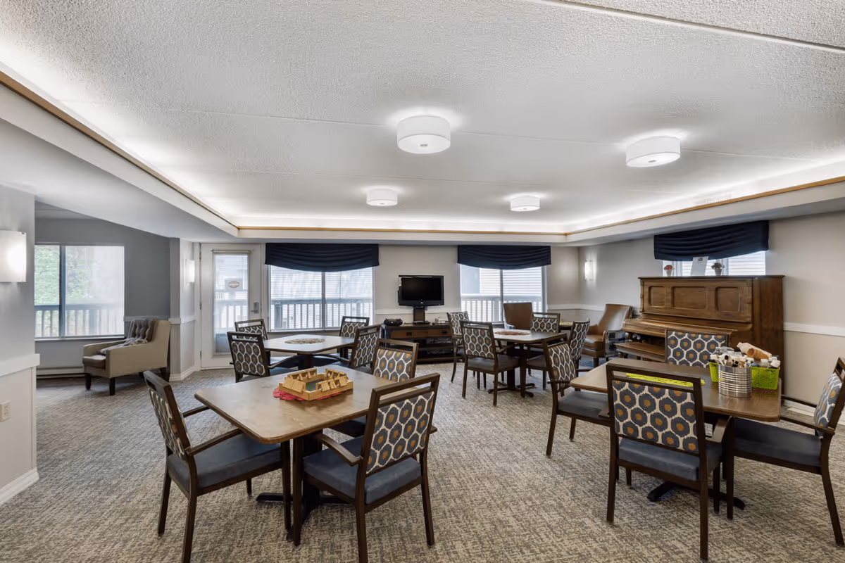 A senior living facility common room with multiple tables and chairs arranged for group activities. The room features a piano against the wall, a television mounted on a stand, and large windows with dark blue valances letting in natural light. The carpeted floor and neutral-colored walls create a cozy atmosphere.
