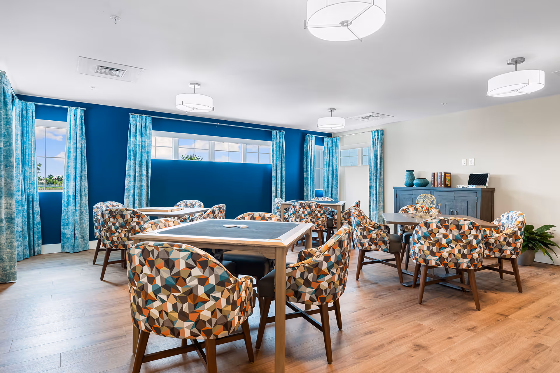 A bright and modern dining room with multiple tables and colorful geometric-patterned chairs. The room features large windows with blue and white curtains, blue accent walls, light wood flooring, and ceiling lights. A dark wooden cabinet with decorative items and books is against one wall.