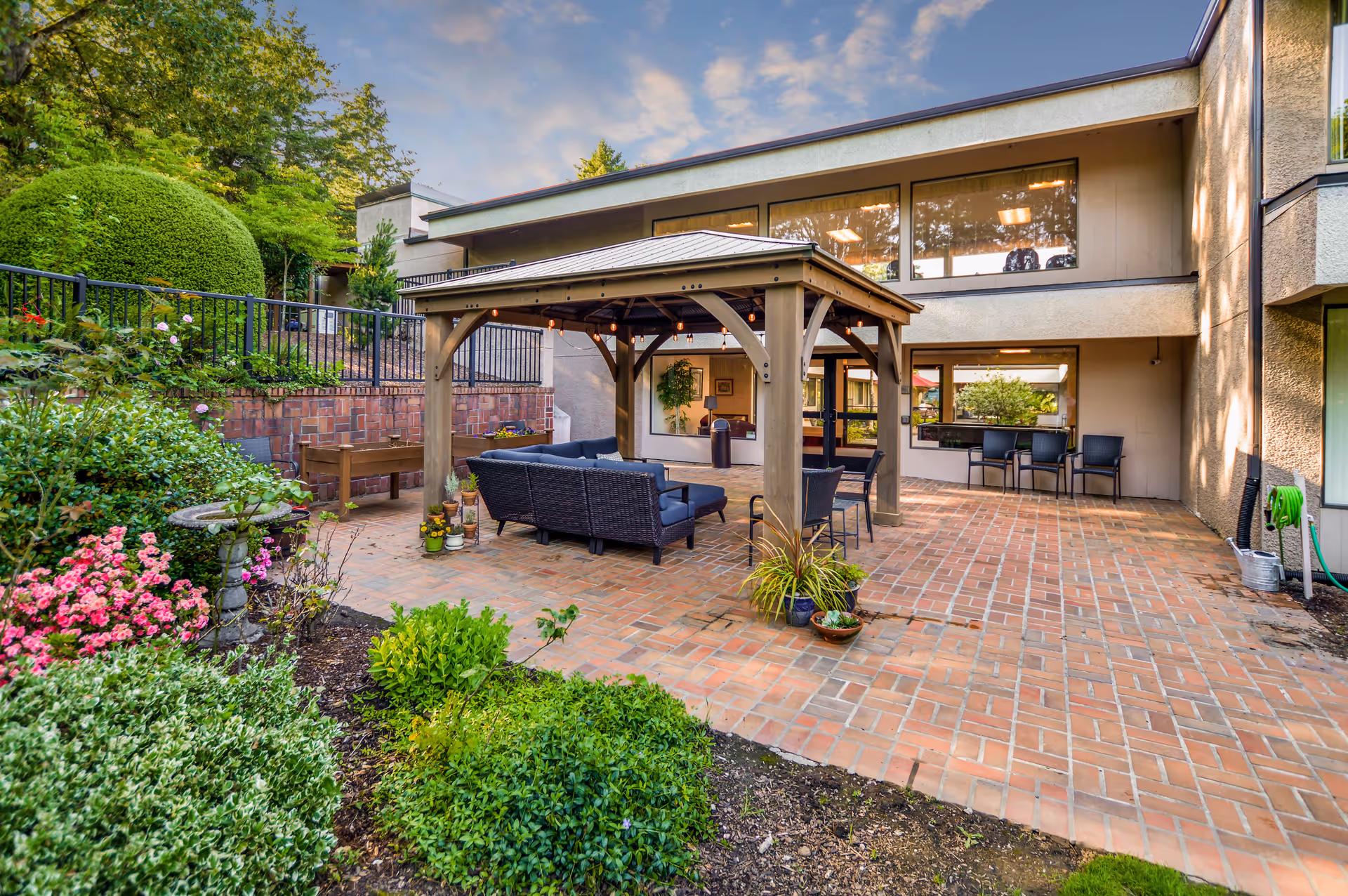 Outdoor patio area at Greenridge Estates Assisted Living featuring a wooden gazebo with seating underneath, surrounded by brick flooring and lush greenery including bushes and flowering plants. The building exterior with large windows is visible in the background under a partly cloudy sky.