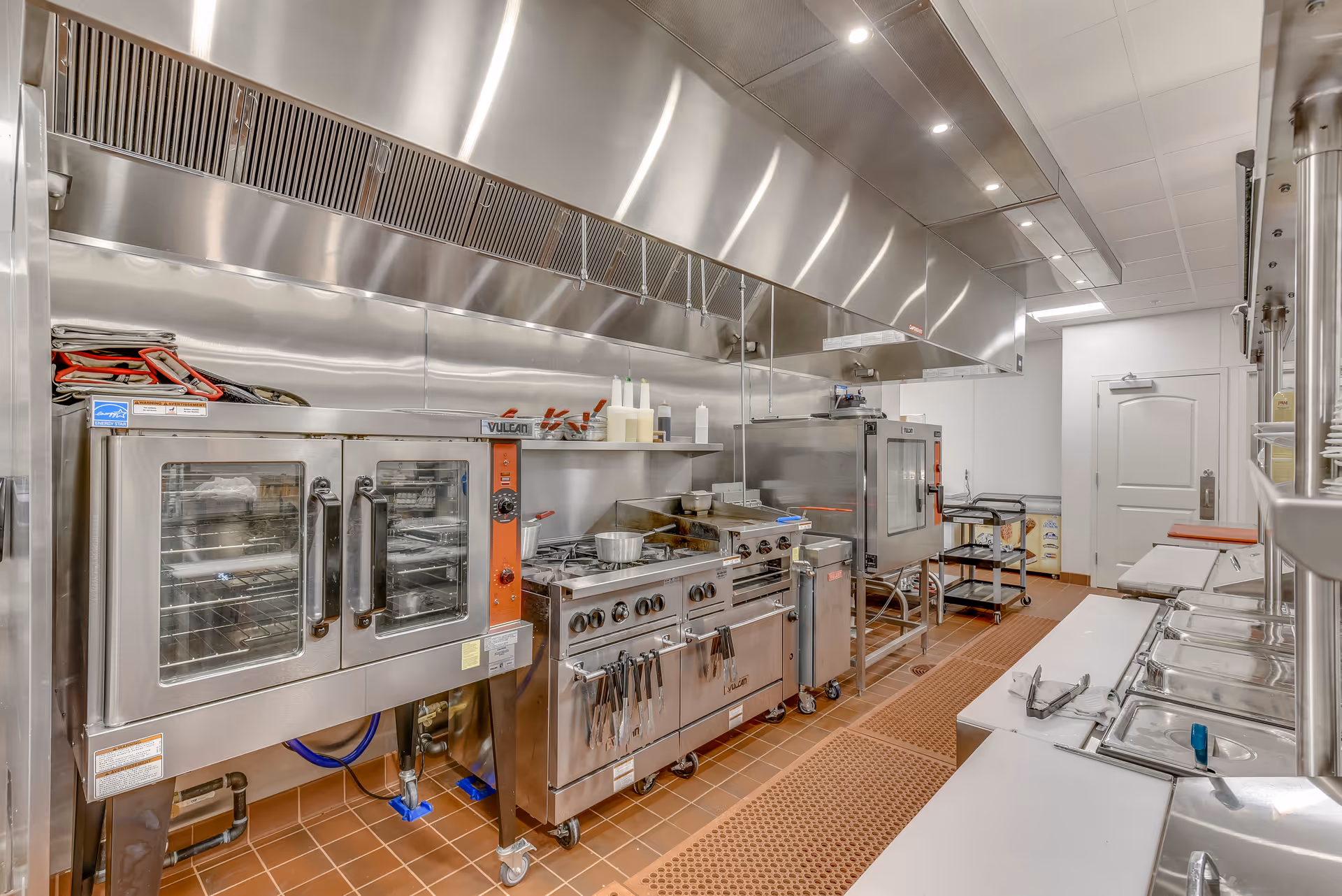 A commercial kitchen with stainless steel appliances including ovens, stovetops, and a large ventilation hood. The kitchen has a clean, organized appearance with various cooking utensils and containers on the counters and shelves. The floor is tiled in a reddish-brown color and there is a white door at the far end of the room.