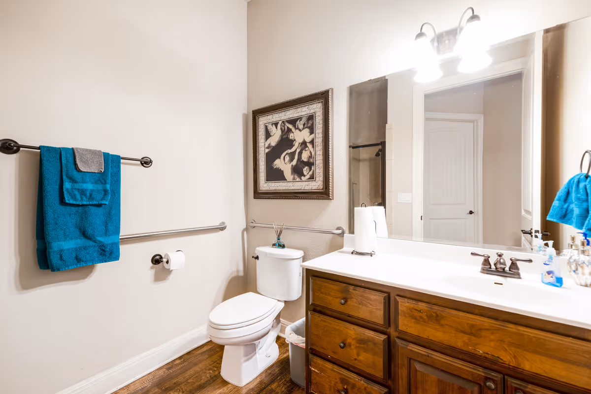 Bright bathroom with a toilet, wooden vanity and sink, large mirror, towel racks with teal towels, and framed artwork on the wall.