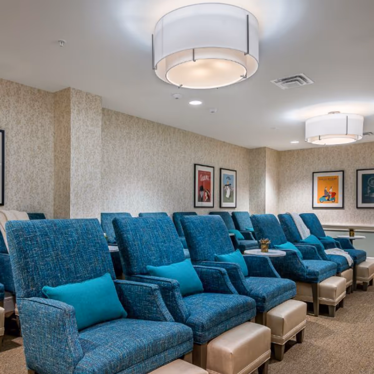 A room with multiple blue upholstered reclining chairs, each paired with a small ottoman and a turquoise pillow. The walls are decorated with framed artwork, and the ceiling has modern circular light fixtures.