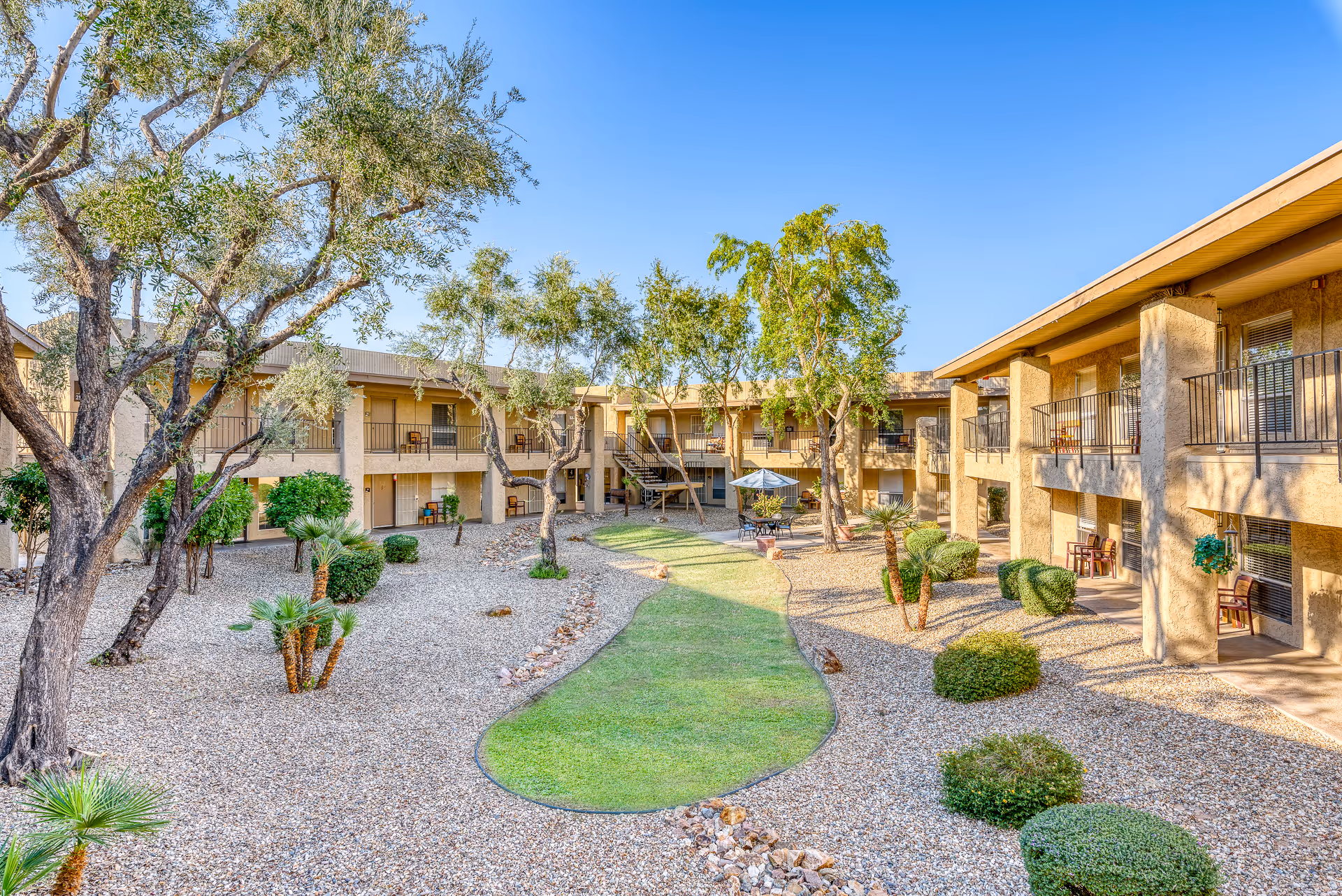 Sunlit two-story courtyard with balconies, gravel landscaping, trees, and a small central lawn at a senior living complex.