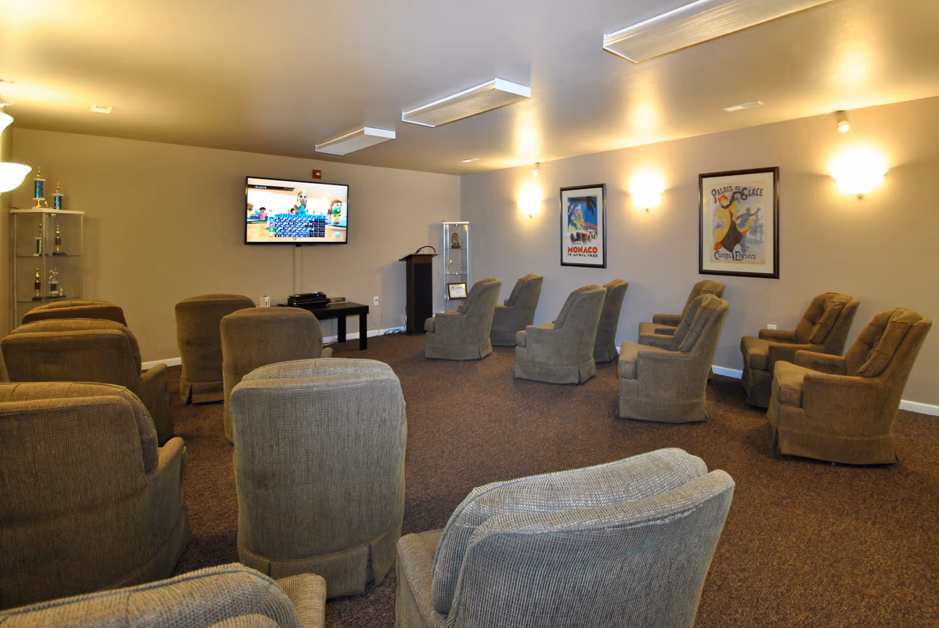 A cozy lounge/media room with rows of upholstered recliner chairs facing a wall-mounted TV and framed posters on the walls.