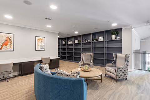 A modern common area with a curved blue sofa, two patterned armchairs, and a round wooden coffee table with a small plant centerpiece. Behind the seating area is a large dark built-in bookshelf with decorative items. To the left, there is a long desk with two office chairs and framed artwork on the wall. The room has light-colored walls and carpeted flooring with recessed ceiling lights.