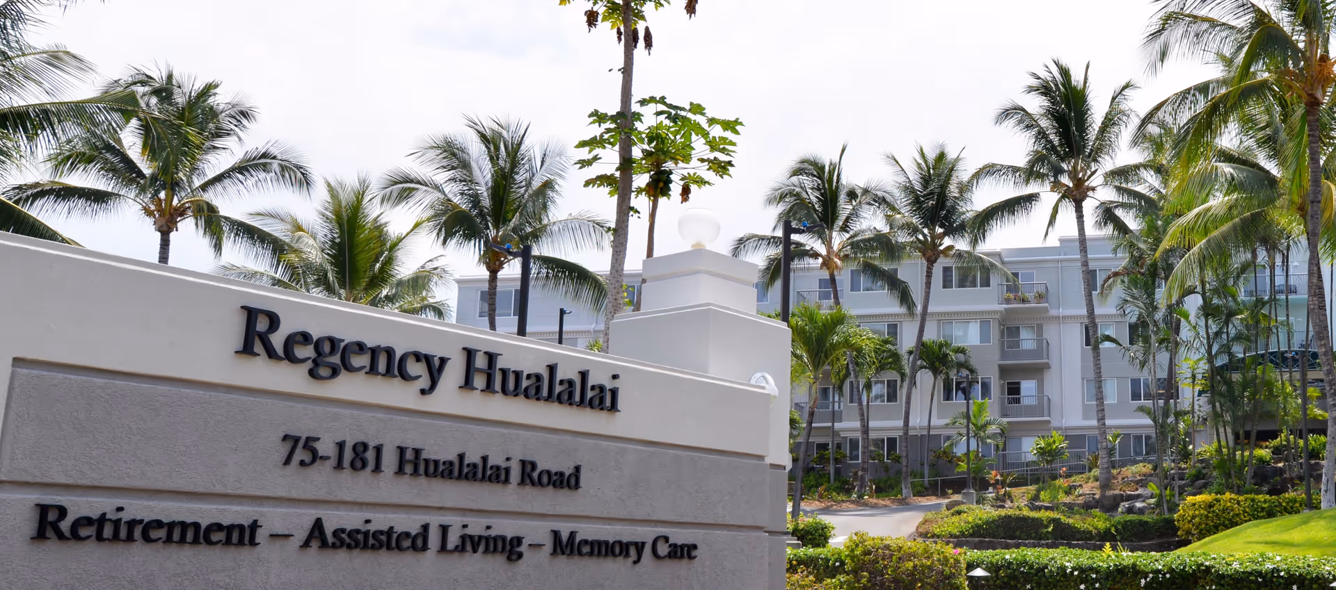 Exterior view of Regency Hualalai senior living facility with a sign displaying the name, address, and services offered. The building is surrounded by palm trees and landscaped greenery under a partly cloudy sky.