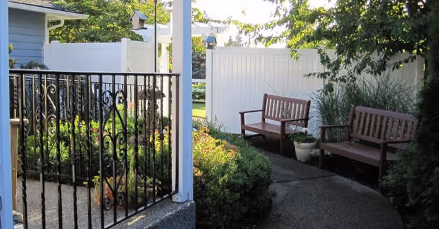 Outdoor garden area with two wooden benches placed along a curved concrete pathway, surrounded by green bushes and plants. A black wrought iron gate is visible on the left side, and a white fence encloses the area. Trees and sunlight are seen in the background.
