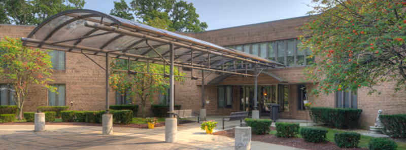 Outdoor view of Robbinswood Northcrest Campus showing a brick building with large windows, a covered walkway with a curved transparent roof, benches, potted plants, and neatly trimmed bushes and trees.