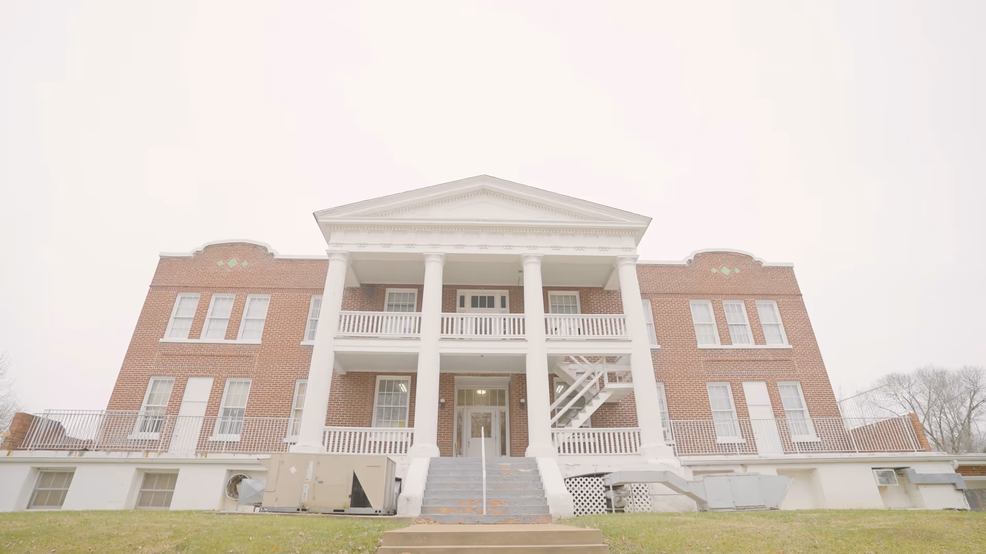 Front exterior view of a large brick building with white columns and a triangular pediment, featuring multiple windows and a central staircase leading to the entrance.