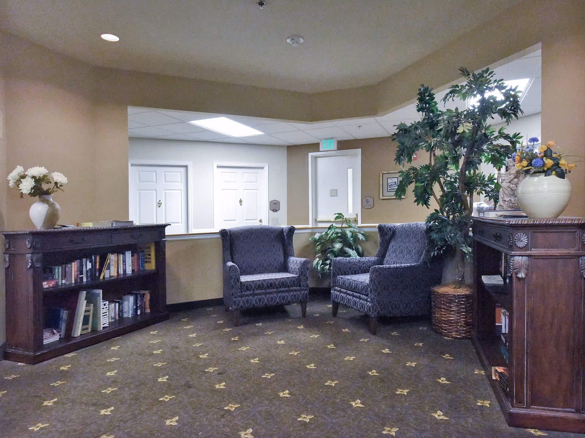 A cozy seating area in a senior living facility with two patterned armchairs, two wooden bookshelves filled with books, potted plants, and floral arrangements. The walls are beige, and there are white doors and an exit sign in the background.