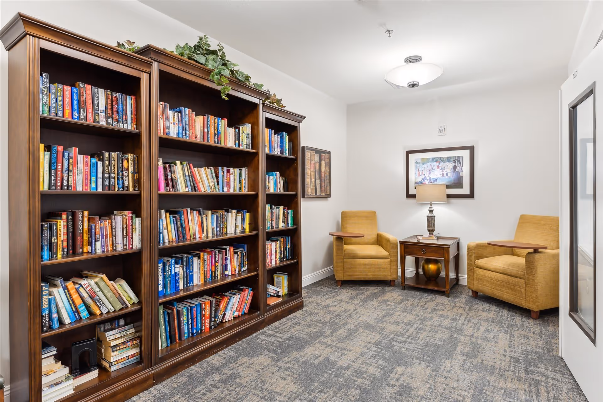 A cozy reading nook in a senior living facility featuring a large wooden bookshelf filled with books, two mustard-yellow armchairs with attached wooden side tables, a small wooden side table with a lamp and decorative vase, and framed artwork on the walls. The room has a patterned carpet and white walls with a ceiling light fixture.