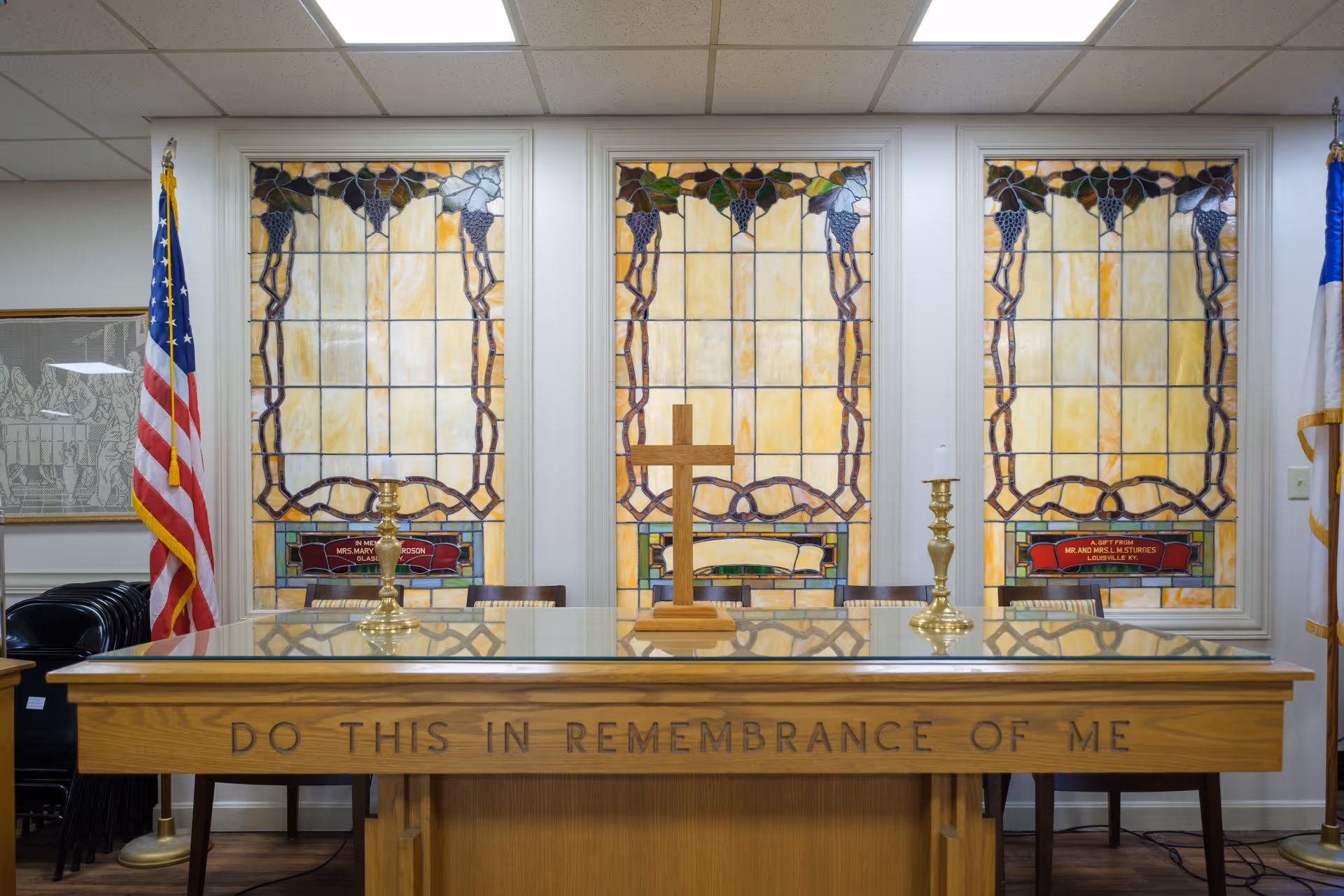 Interior view of a chapel or memorial room with a wooden altar inscribed with 'DO THIS IN REMEMBRANCE OF ME'. Behind the altar are three stained glass windows with grapevine designs. On the altar are two brass candlesticks with white candles and a wooden cross in the center. An American flag is positioned to the left and another flag to the right.