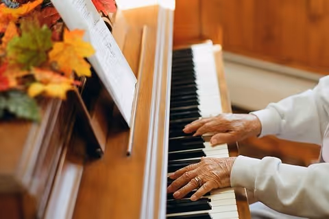 Close-up of an elderly person's hands playing a wooden piano with sheet music and autumn-themed decorations on top of the piano.
