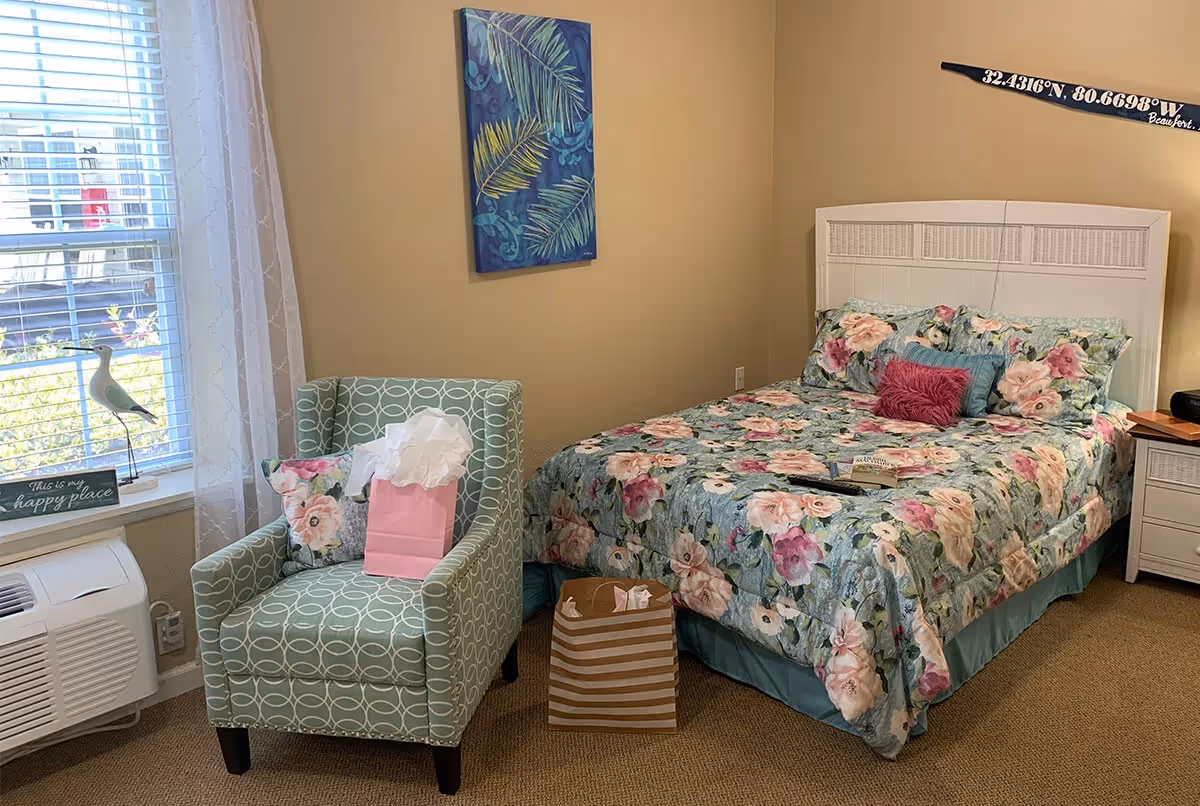 A cozy bedroom with a floral-patterned bedspread on a white bed frame, a green patterned armchair with a floral pillow and a pink gift bag on it, a window with white curtains, a small nightstand with a lamp, and a decorative wall art featuring palm leaves. There is also a striped gift bag on the floor next to the bed.