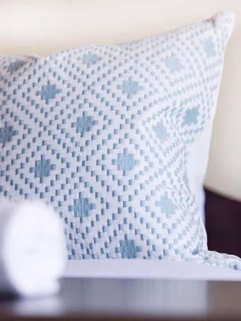 Close-up of a decorative pillow with a geometric pattern in light blue and white, placed on a bed with white sheets.