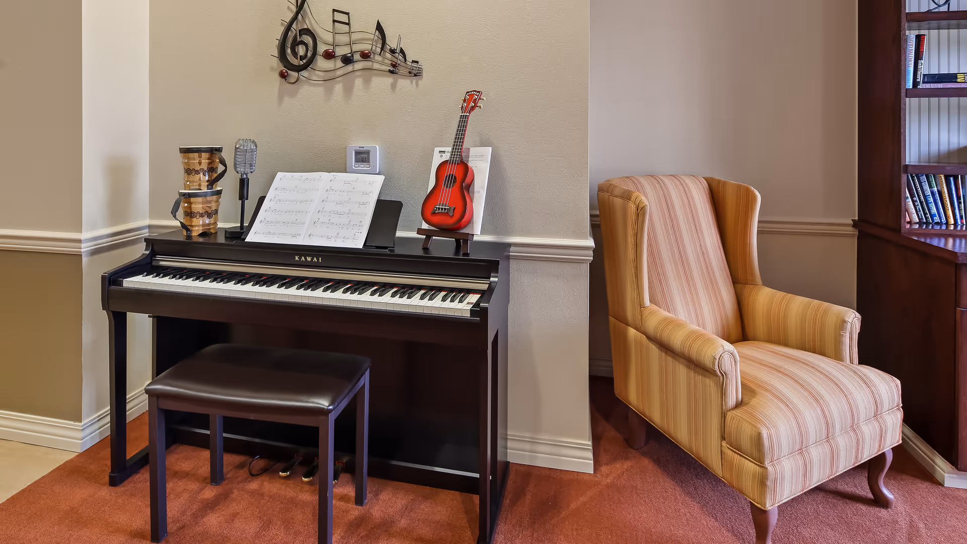 A cozy corner featuring a black Kawai digital piano with sheet music, a small red ukulele on a stand, two wooden percussion drums, and a vintage-style microphone. Next to the piano is a comfortable striped armchair in warm tones, and a wooden bookshelf filled with books is partially visible on the right.