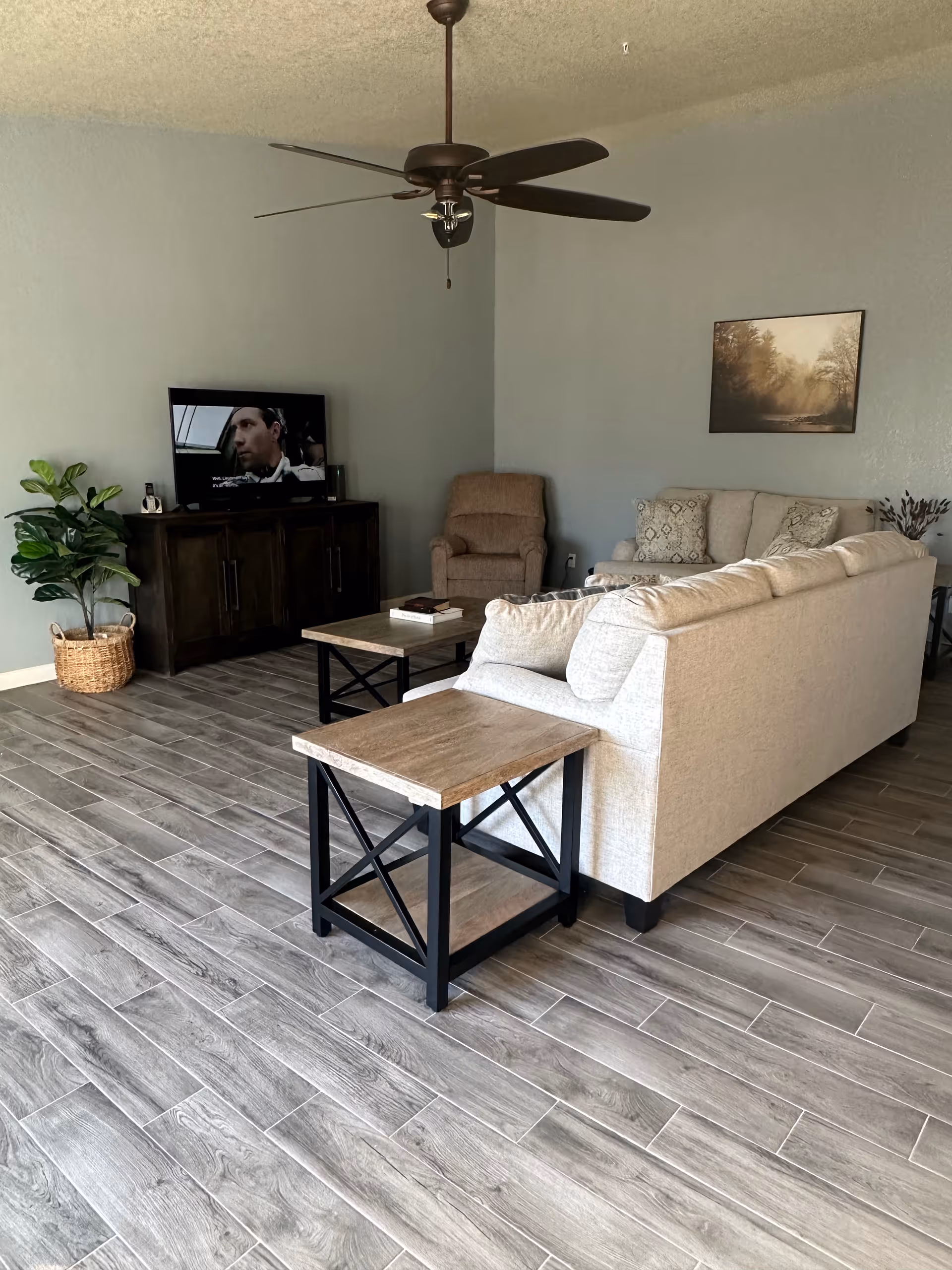 A cozy living room with a beige sectional sofa, a matching armchair, and a wooden coffee table and side table with black metal frames. A flat-screen TV is placed on a dark wooden TV stand against a light gray wall. A potted plant in a woven basket is next to the TV stand. The floor has gray wood-look tiles, and a ceiling fan with dark blades hangs from the ceiling. A framed landscape picture is on the wall above the sofa.