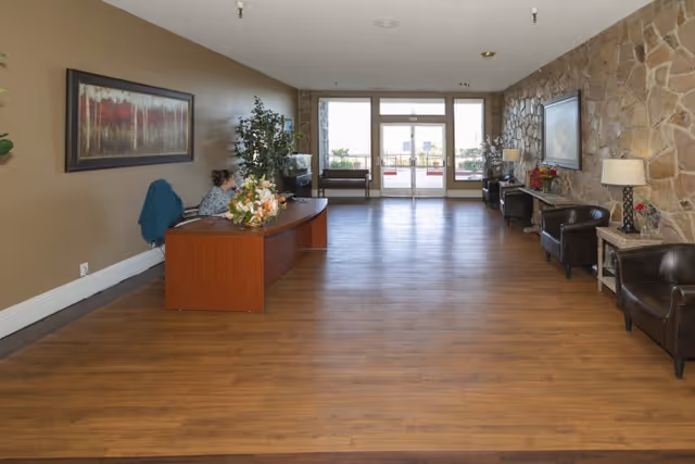 A spacious reception area with wooden flooring and a stone accent wall on the right. There is a wooden reception desk with a person seated behind it on the left side, decorated with a flower arrangement. The room has several leather chairs along the right wall, two framed paintings, and lamps on side tables. Large glass doors at the far end lead outside, allowing natural light into the room.