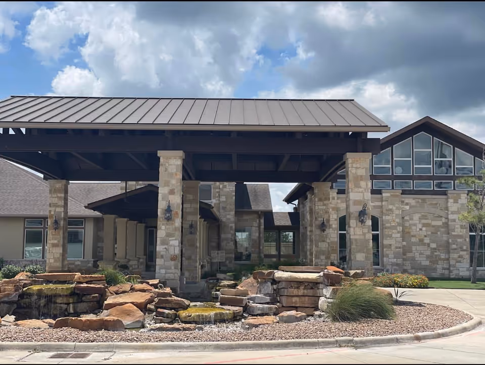 Front exterior view of North Twin Pines Nursing Home featuring a covered entrance with stone pillars, a rock water feature in front, and a building with large windows under a partly cloudy sky.