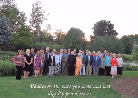 A large group of people standing together outdoors on a grassy area with trees and bushes in the background during sunset. They are posing for a group photo and dressed in a variety of formal and semi-formal attire. Text at the bottom reads: 'Meadows, the care you need and the dignity you deserve.'