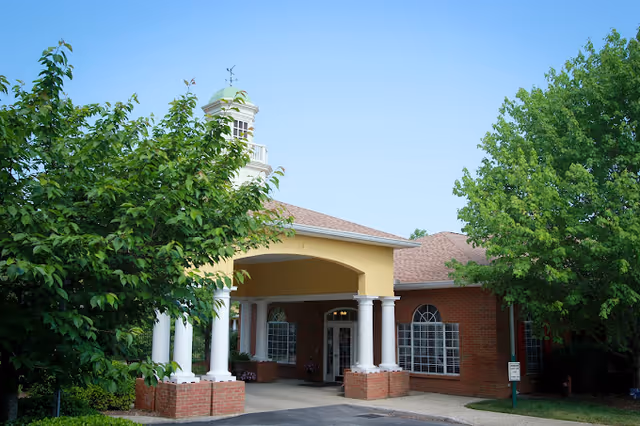 Exterior view of a senior living facility building with a covered entrance supported by white columns, surrounded by green trees and a clear blue sky.