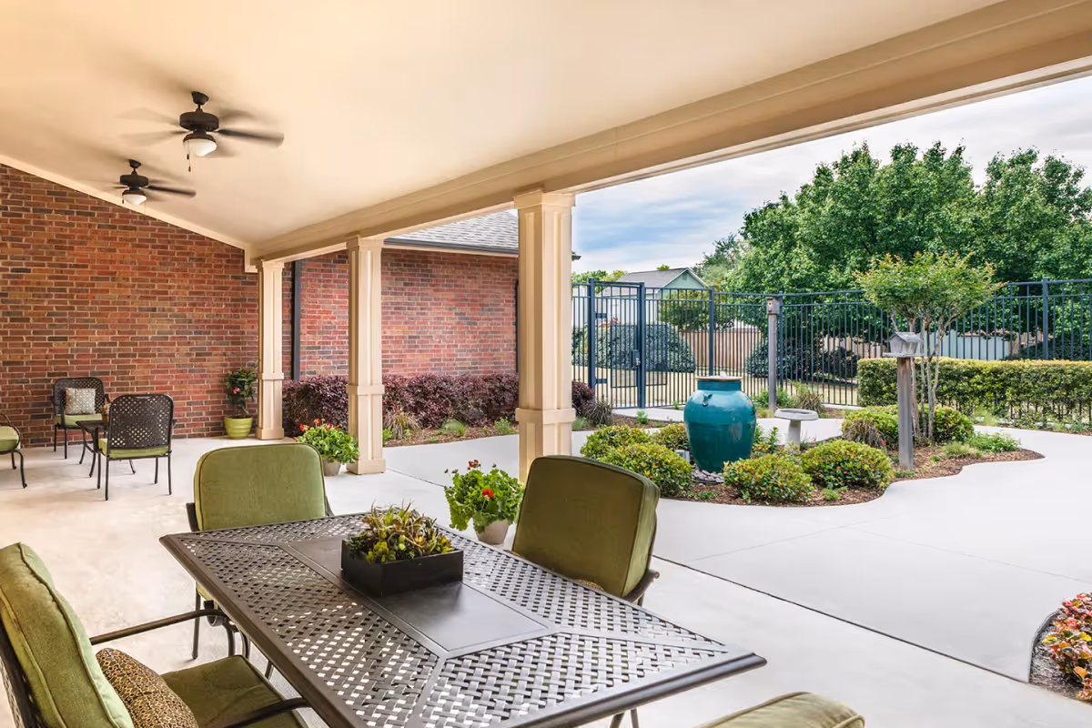 Covered outdoor patio area with green cushioned chairs and a metal table, ceiling fans, and a view of a landscaped garden with bushes, a large blue ceramic pot, and a black metal fence in the background.