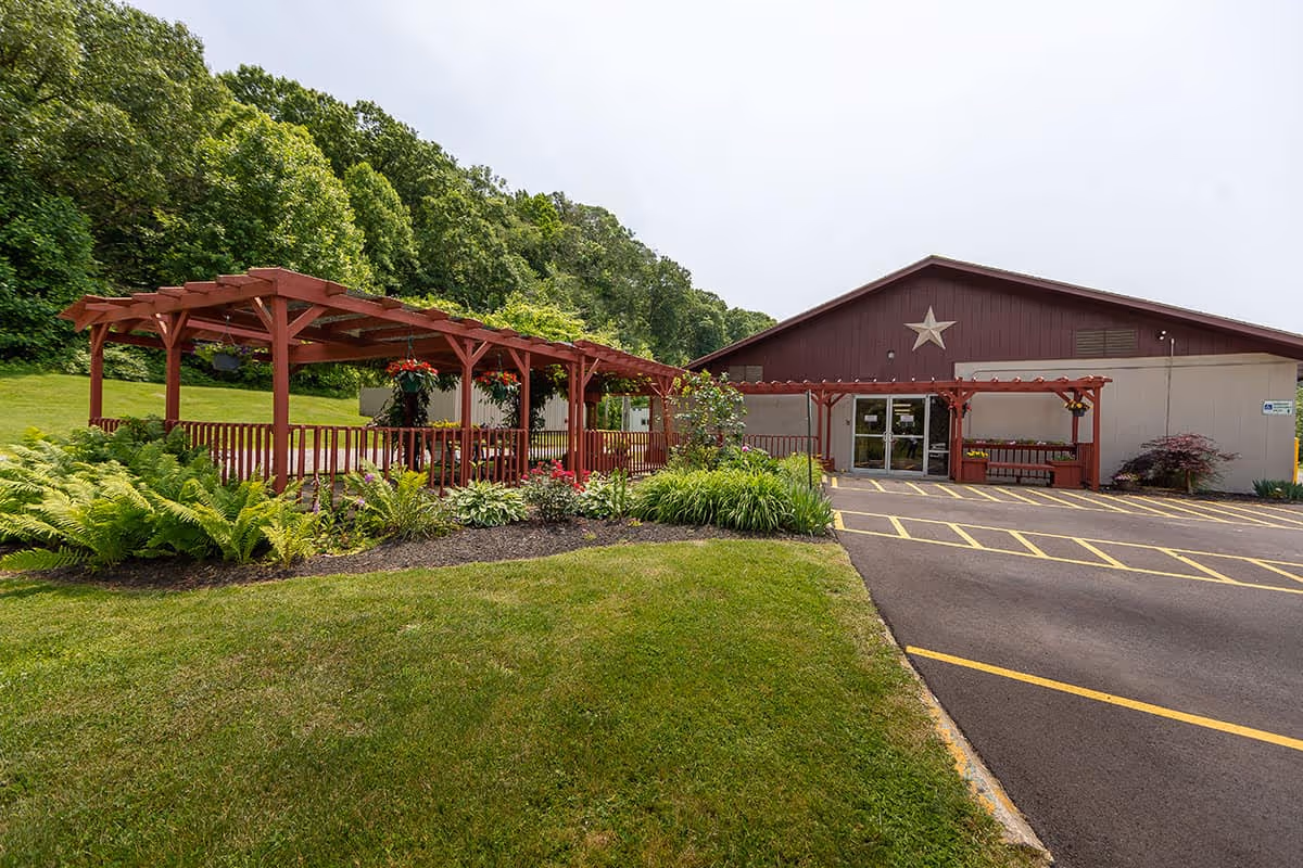 Front exterior of a single-story red building with a large star above the entrance, a red pergola-covered walkway, parking spaces, and landscaped lawn.