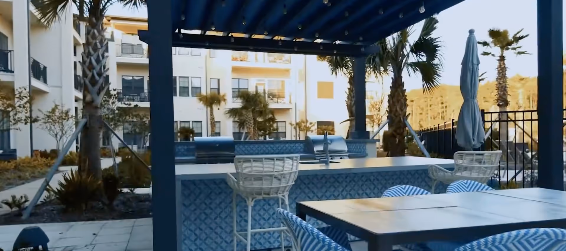 Outdoor seating area with a blue pergola, bar counter with two grills, white wicker bar stools, and a table with blue patterned chairs. Palm trees and a multi-story building are visible in the background.