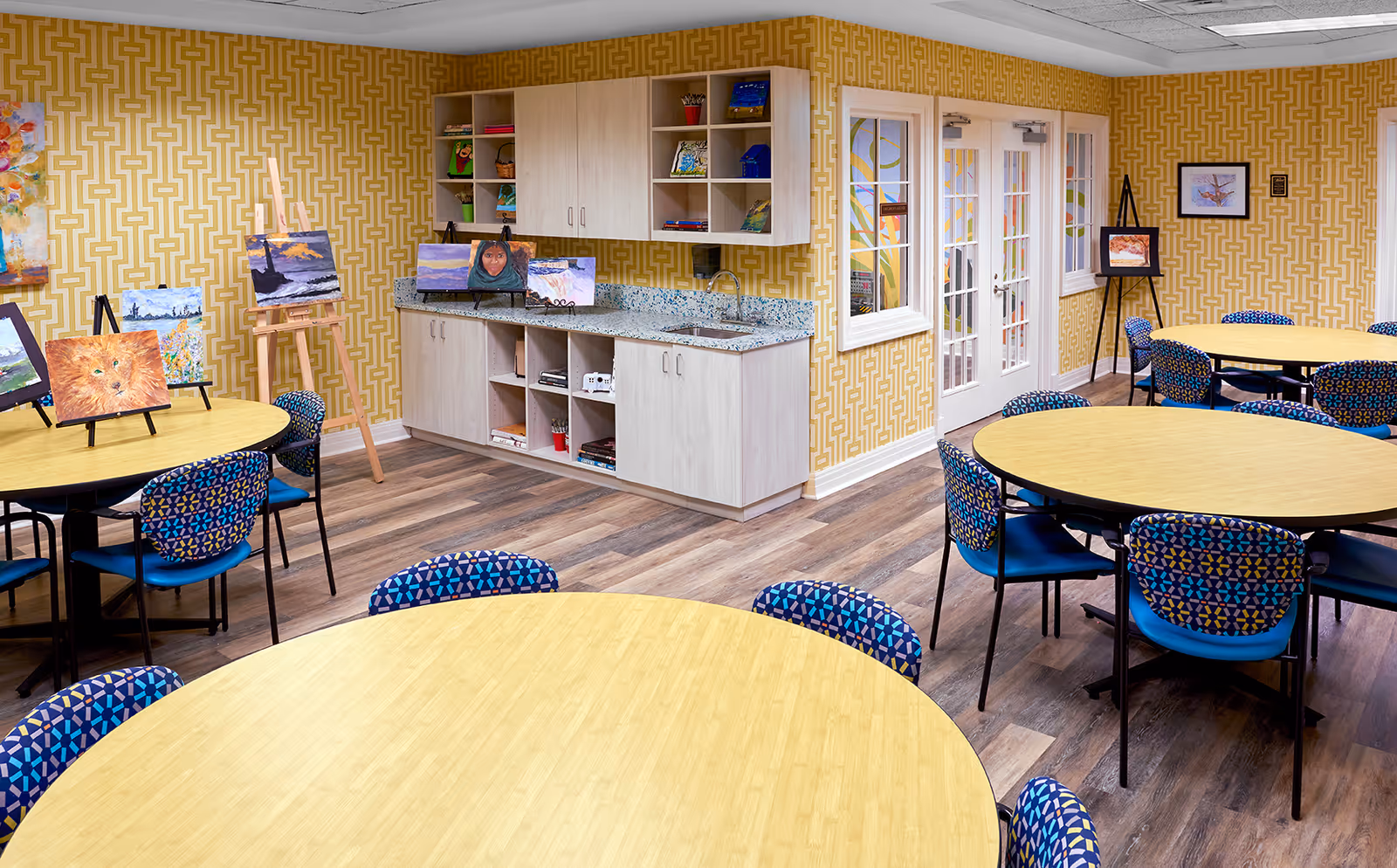 A bright and colorful activity room with round wooden tables surrounded by blue patterned chairs. The walls have a yellow geometric pattern wallpaper. Several paintings and art supplies are displayed on easels and shelves along the walls. There is a countertop with cabinets and a sink, and double glass doors leading to another room.