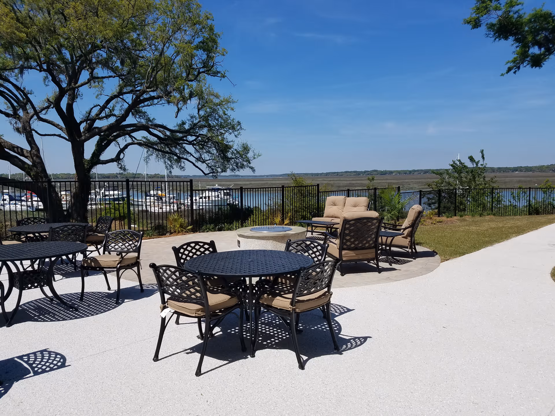 Outdoor patio area with black metal tables and cushioned chairs arranged around a fire pit, overlooking a fenced marina with boats and a large tree providing shade on a sunny day.
