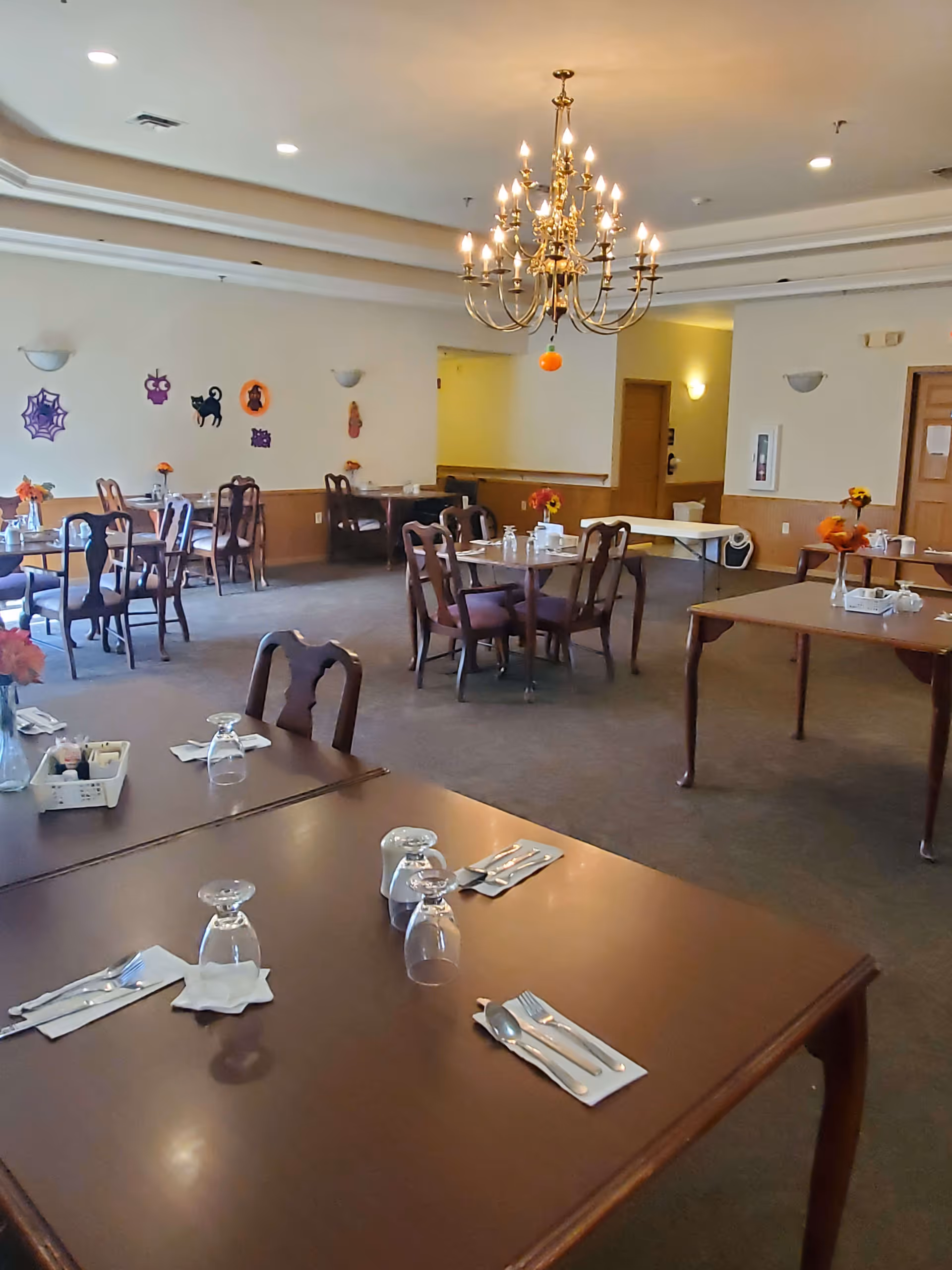 A dining room with multiple wooden tables and chairs arranged neatly. Each table is set with upside-down glasses, napkins, and cutlery. The room is decorated with small flower vases on the tables and Halloween-themed wall decorations including a spider web, black cat, and pumpkins. A large chandelier hangs from the ceiling providing warm lighting.