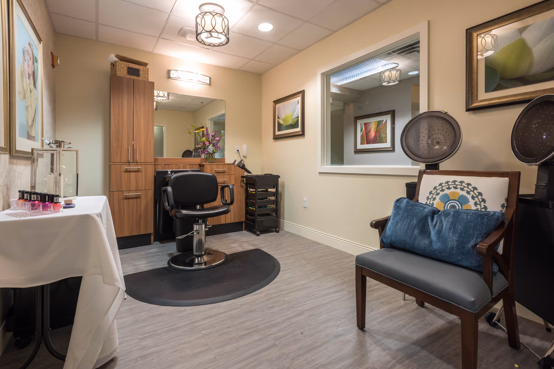 Small salon interior with a styling chair in front of a mirror and wooden cabinets, hooded hair dryers, and a waiting chair with pillows.