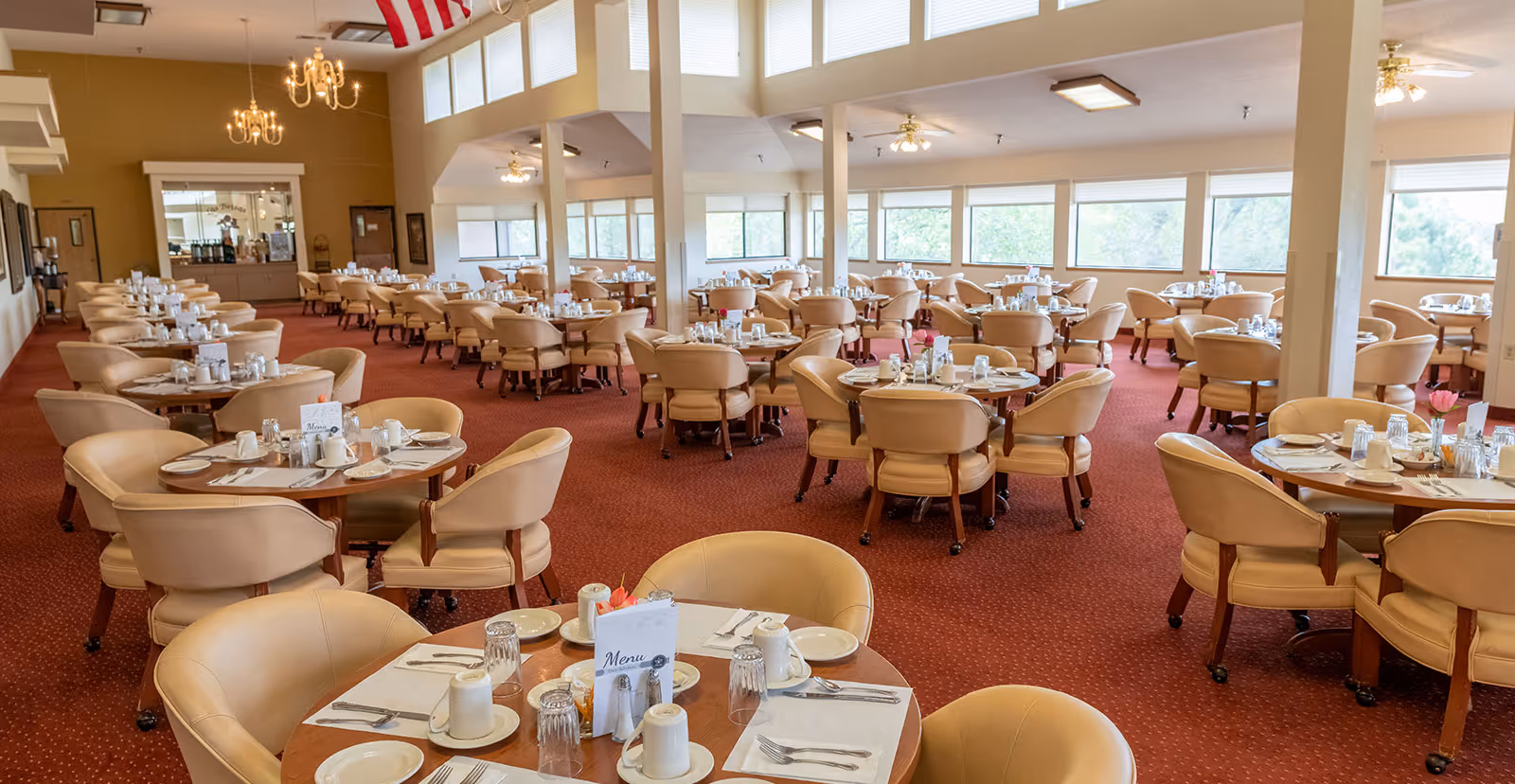 A spacious dining room with multiple round tables set with plates, cups, utensils, and menus. The room has beige upholstered chairs, red carpet flooring, large windows letting in natural light, and chandeliers hanging from the ceiling.