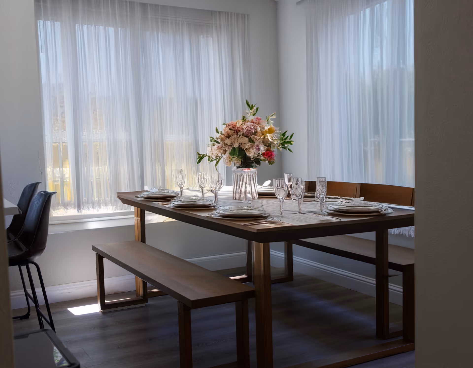 Sunlit dining area with a wooden table set with plates, glassware, and a floral centerpiece, flanked by benches and sheer curtains.