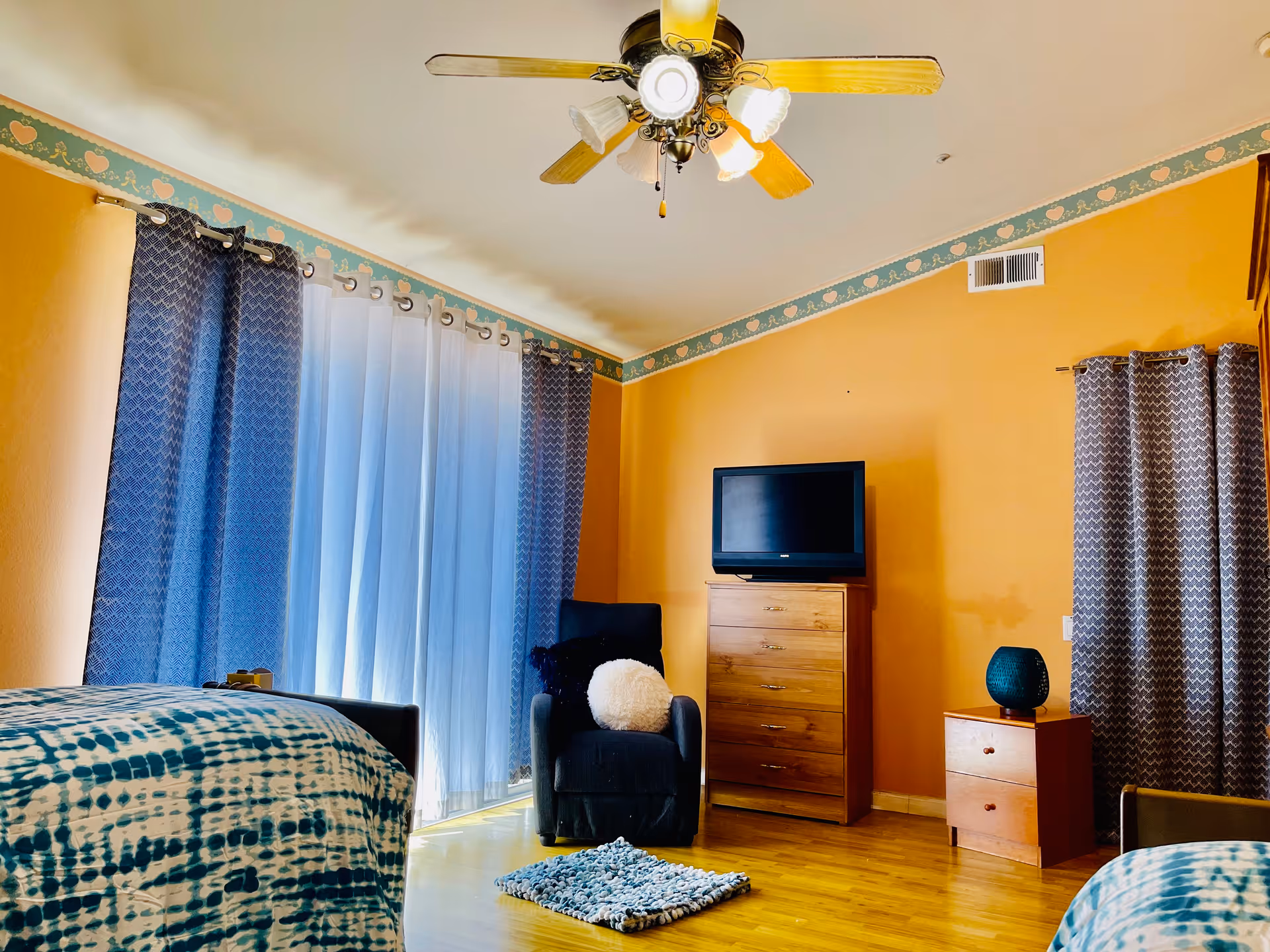 Bright bedroom with two beds, a dresser and TV, an armchair, and blue patterned curtains.