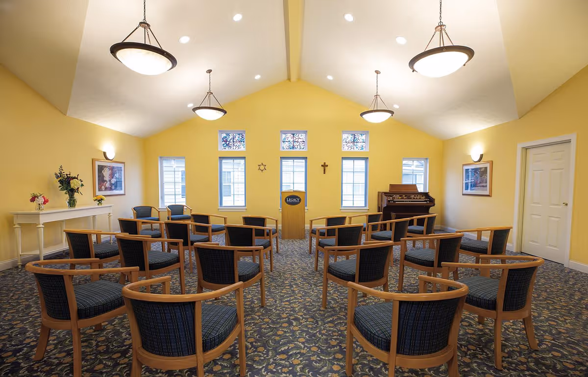Interior of a small chapel or meeting room with yellow walls, a high vaulted ceiling with hanging lights, rows of wooden chairs with blue cushions arranged facing a wooden podium. The room has several windows with stained glass at the top, a cross and a Star of David on the wall behind the podium, framed pictures on the walls, a white console table with flower arrangements, and an organ in the corner.
