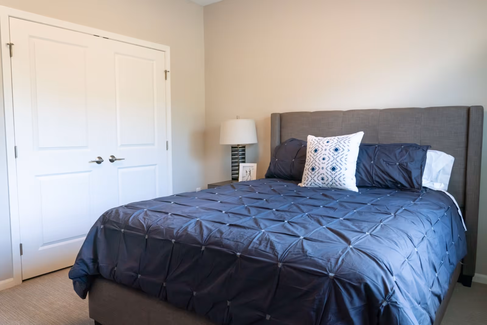 A bedroom with a neatly made bed featuring a dark gray comforter and three pillows, one with a white and blue geometric pattern. There is a nightstand with a lamp and a small framed picture next to the bed. The walls are light beige, and there is a white double-door closet on the left side of the room.