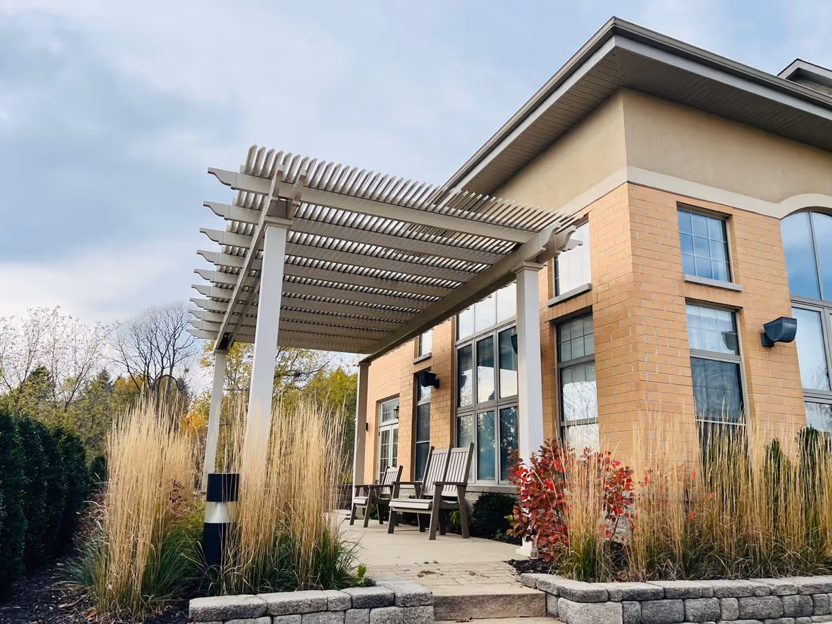 Exterior view of a brick assisted living building with a pergola-covered patio, chairs, and ornamental grasses.