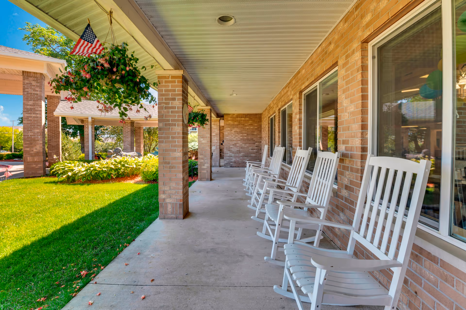 Covered front porch with a row of white rocking chairs along a brick building, hanging plants and an American flag overlooking a green lawn.