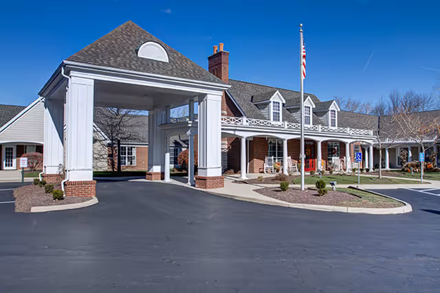 Exterior view of a senior living facility with a covered entrance, brick and white siding building, American flag on a flagpole, and a paved driveway with parking spaces.