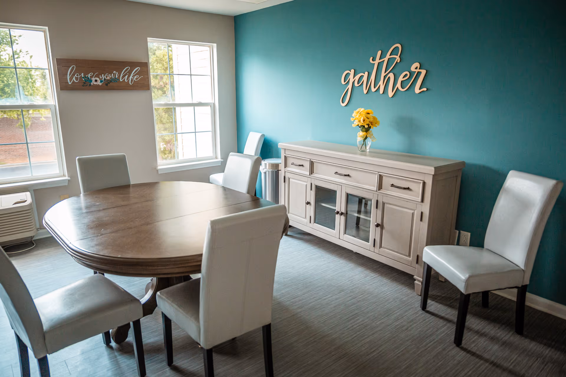 A dining room with a wooden oval table surrounded by six white cushioned chairs. The room has two large windows letting in natural light, a teal accent wall with the word 'gather' in wooden letters, and a light-colored sideboard with a vase of yellow flowers on top. A wall decoration reads 'love your life'.